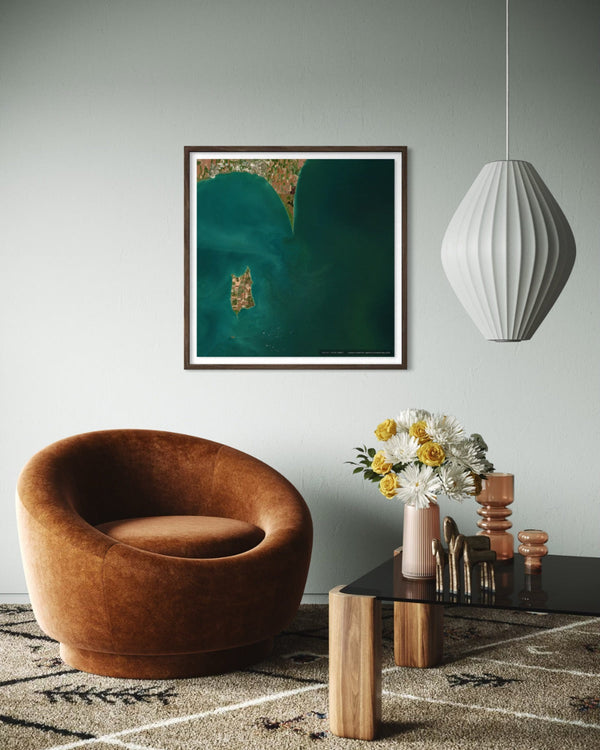A framed abstract art print featuring deep teal and green tones with gold accents, displayed on a light gray wall in a modern living room setting with a brown velvet armchair, a black coffee table, and a white pendant light.