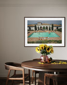 A framed art print depicting a Mediterranean-style villa with a turquoise swimming pool, displayed on a beige wall above a wooden dining table with chairs and a vase of sunflowers.