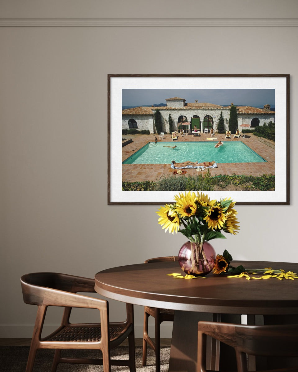 A framed art print depicting a Mediterranean-style villa with a turquoise swimming pool, displayed on a beige wall above a wooden dining table with chairs and a vase of sunflowers.