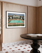 A framed art print depicting a luxurious outdoor poolside scene with a villa and lounge chairs, displayed on a light wood-paneled wall in a modern interior setting.