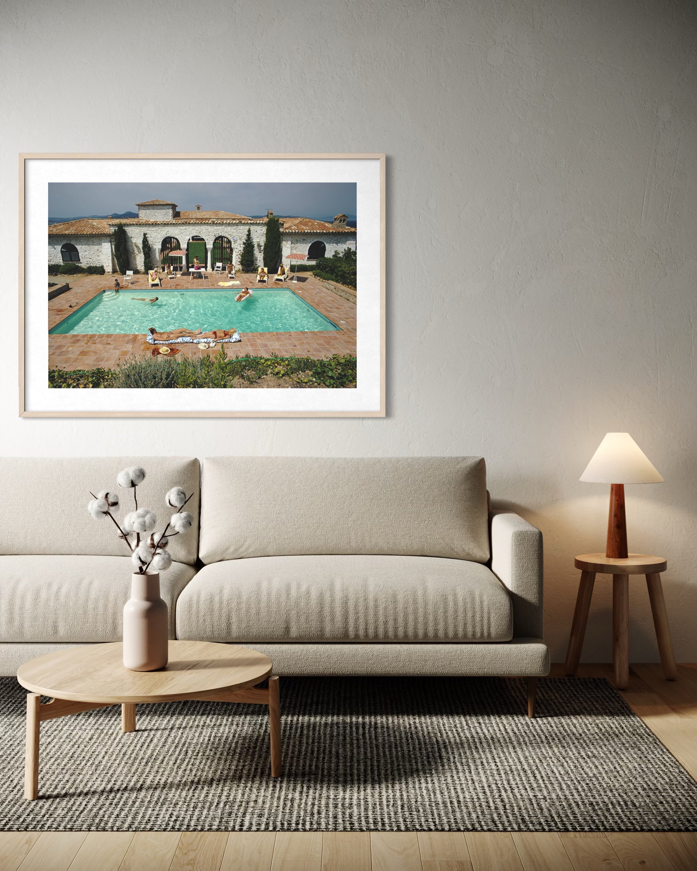 A framed art print depicting a luxurious villa with a turquoise swimming pool, displayed on a light gray wall above a modern beige sofa in a styled living room.