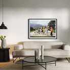 A framed art print depicting a group of people playing with a pool table and skateboards in front of mountains, displayed on a light gray wall above a modern beige sofa in a styled living room.
