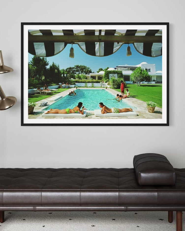 Framed artwork of people by a pool on a wall above a brown leather daybed.