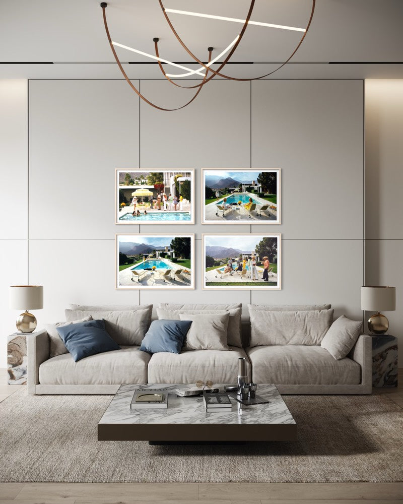 A modern living room with a light beige sofa, marble coffee table, and a wall-mounted set of four framed photographs above the couch. The photos depict outdoor poolside and mountain scenes. The Picturalist Wall Art and Photography for Designed Interiors. Slim Aarons Collection, Poolside. Getty Images Gallery. 