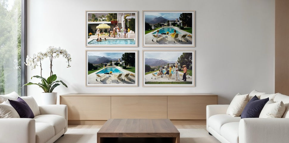 A modern living room with two white sofas and a wooden coffee table, featuring a wall display of four framed square photographs showing poolside resort scenes. The Picturalist Wall Art and Photography for Designed Interiors. Slim Aarons Collection, Poolside. Getty Images Gallery. 
