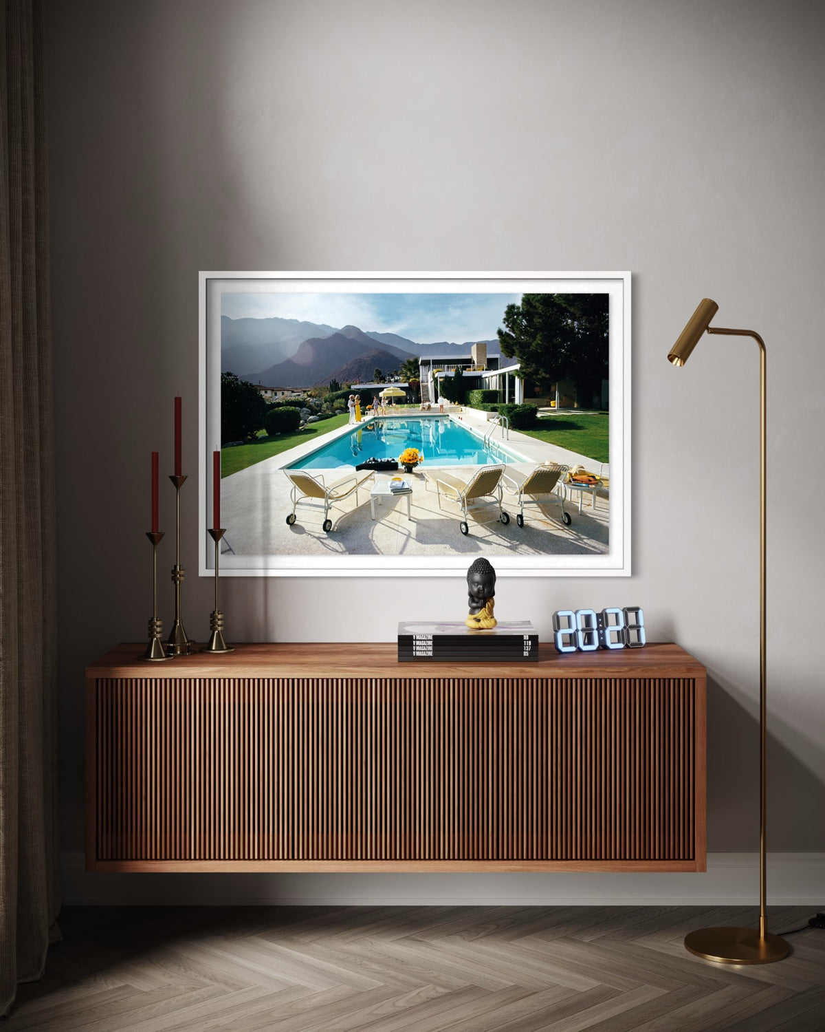 A framed art print depicting a luxurious outdoor poolside scene with mountains in the background, displayed on a gray wall above a wooden console table in a modern living room setting.