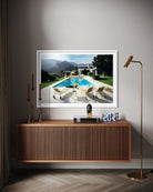 A framed art print depicting a luxurious outdoor poolside scene with mountains in the background, displayed on a gray wall above a wooden console table in a modern living room setting.