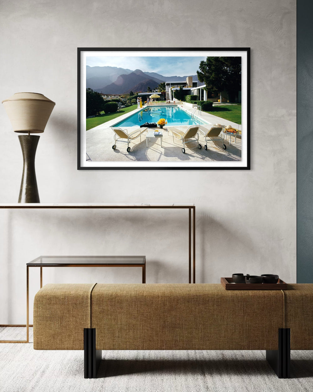 A framed art print depicting a luxurious outdoor poolside scene with mountains in the background, displayed on a textured light gray wall above a modern console table and bench.