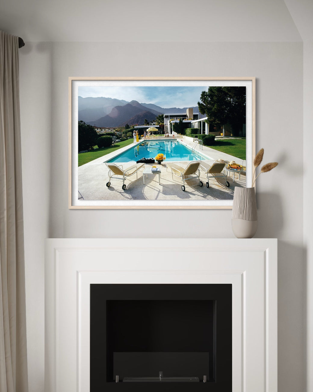 A framed art print depicting a luxurious outdoor poolside scene with mountains in the background, displayed on a white wall above a fireplace mantel.