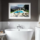 A framed art print depicting a luxurious outdoor poolside scene with lounge chairs, mountains in the background, and a modern house, displayed on a dark gray wall above a white tiled bathroom wall and freestanding bathtub.