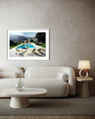 A framed art print depicting a luxurious outdoor poolside scene with mountains in the background, displayed on a textured beige wall above a modern cream-colored sofa in a styled living room.