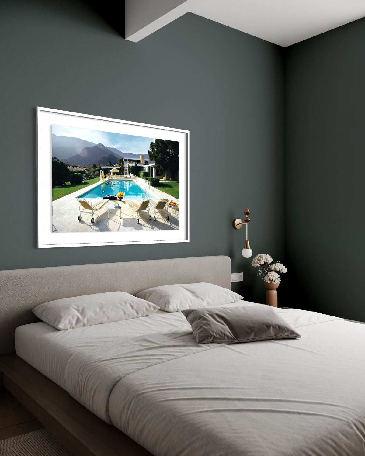 A framed art print depicting a luxurious outdoor poolside scene with mountains in the background, styled on a dark gray wall above a modern bed in a bedroom setting.