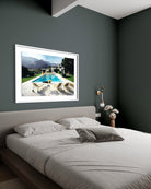 A framed art print depicting a luxurious outdoor poolside scene with mountains in the background, styled on a dark gray wall above a modern bed in a bedroom setting.