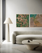 A modern living room scene featuring two framed landscape art prints hanging above a curved light beige sofa. The left print shows green and brown patchwork fields, and the right print shows a winding river or canal in earthy green, brown, and gold tones.