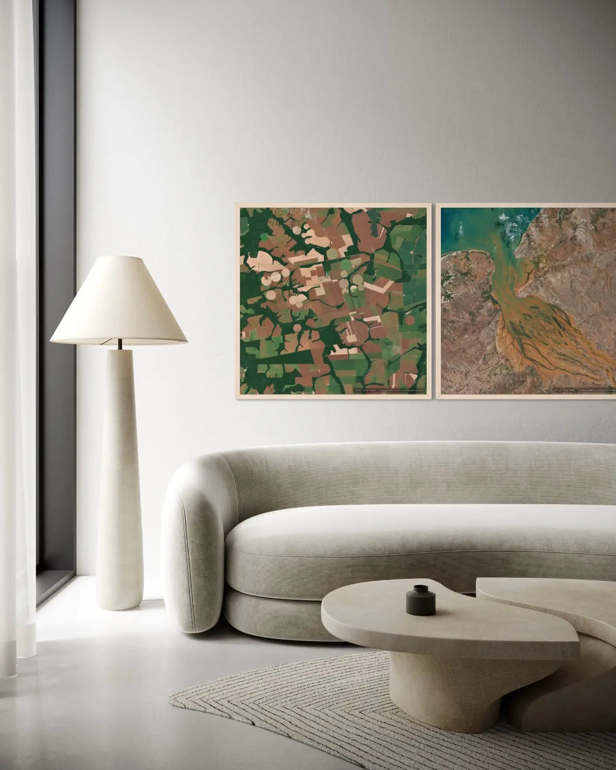 A modern living room scene featuring two framed landscape art prints hanging above a curved light beige sofa. The left print shows green and brown patchwork fields, and the right print shows a winding river or canal in earthy green, brown, and gold tones.