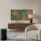 A styled interior scene showing two framed abstract landscape art prints mounted on a beige wall above a dark wood sideboard. The room includes a cream boucle armchair, a black marble side table, a lamp, and a patterned rug.