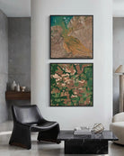 Two framed abstract landscape art prints are displayed vertically on a white wall in a modern living room setting. The top print features a golden-brown bird over green and brown terrain, and the bottom print shows a patchwork of green, brown, and beige fields.