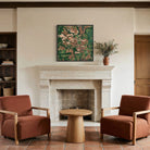 An abstract landscape painting in green, brown, and beige tones hangs on a cream wall above a stone fireplace in a rustic living room setting with two rust-colored armchairs and a wooden side table.