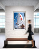 A framed vertical art print of a sailboat scene with white sails and blue sky, displayed on a white brick wall above a wooden bench in a modern office space. A woman in a black dress walks past the frame on the right.