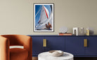 A framed vertical art print of a sailboat on blue water, displayed on a beige wall above a navy blue cabinet in a modern living room setting.