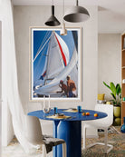A framed vertical art print depicting a sailboat scene with two people on the deck, hanging on a beige wall above a blue dining table in a modern room setting.