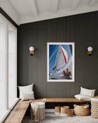 A framed vertical art print of a sailboat on the wall in a modern, nautical-themed room with a dark wood-paneled wall, wooden bench, and woven baskets.