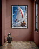 A framed vertical art print of a sailboat scene with white sails, blue sky, and two people on deck, displayed on a dusty rose wall above a dark vase on a wood floor.
