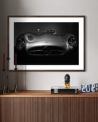 A framed black and white art print of a Mercedes-Benz 300 SL Roadster car, displayed on a light gray wall above a wooden console table with decorative items.