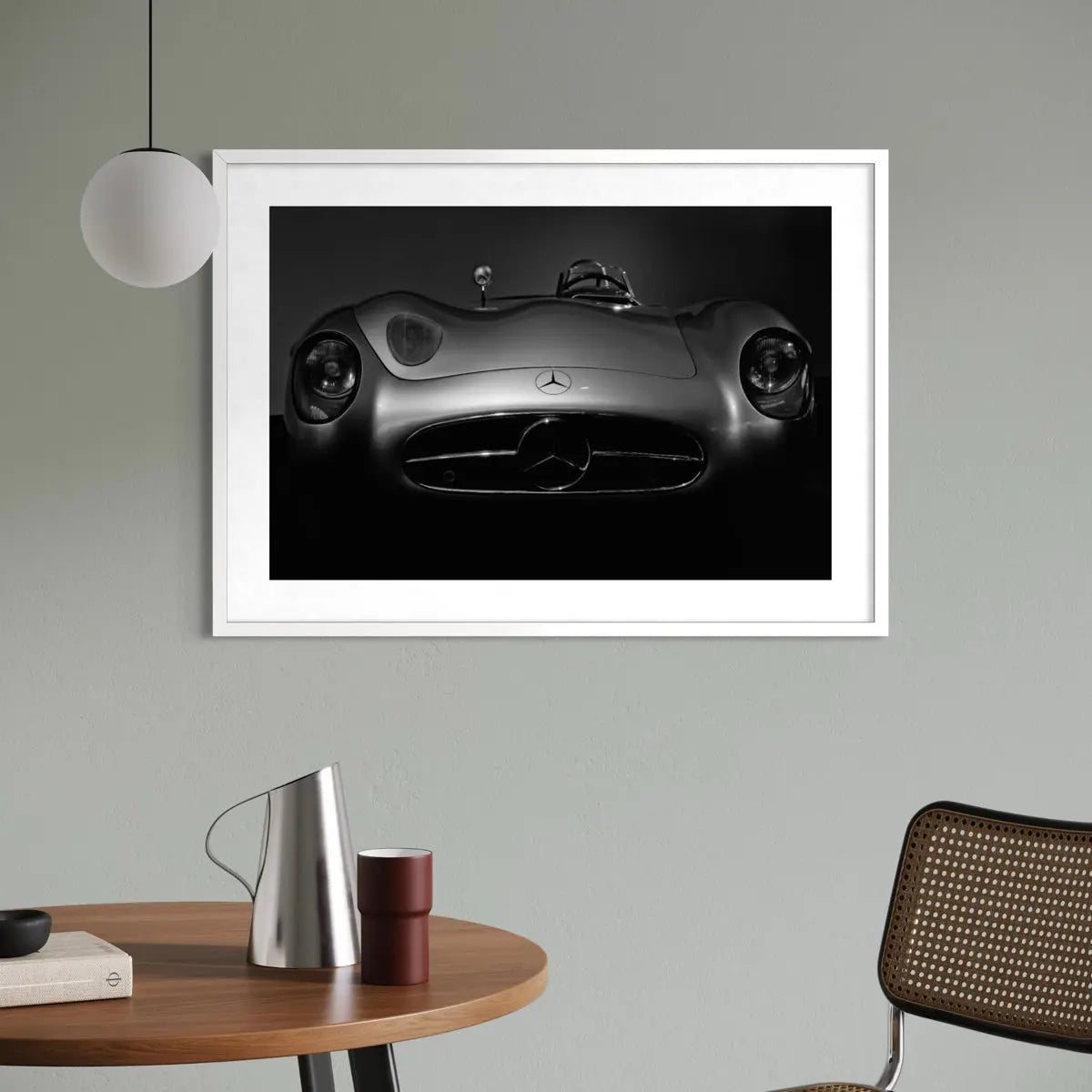 A framed black and white art print of a Mercedes-Benz sports car, displayed on a light gray wall in a modern room setting with a wooden table, lamp, and chair.