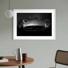 A framed black and white art print of a Mercedes-Benz sports car, displayed on a light gray wall in a modern room setting with a wooden table, lamp, and chair.