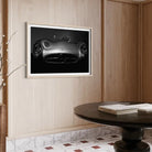 A framed black and white art print of a Mercedes-Benz sports car hanging on a light wood-paneled wall above a dark round table with books.
