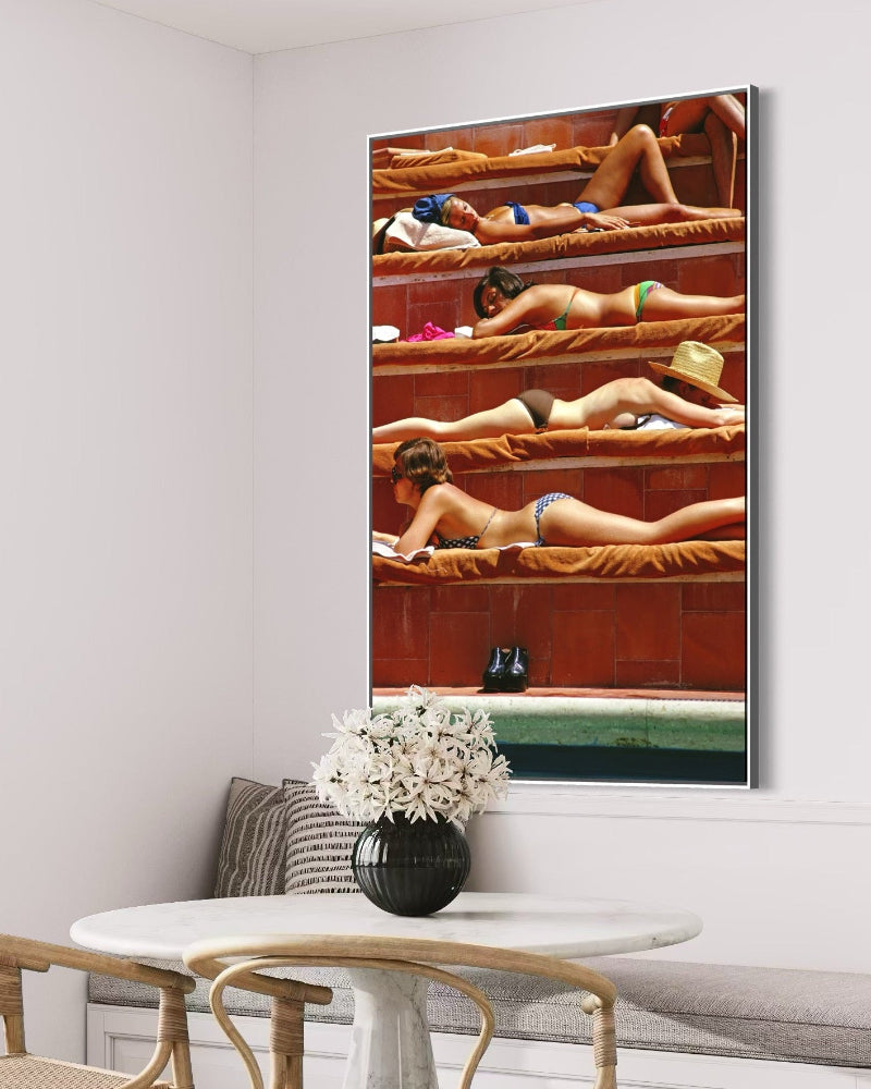 Framed artwork of people in a hot tub on a wall above a table with a vase and flowers.