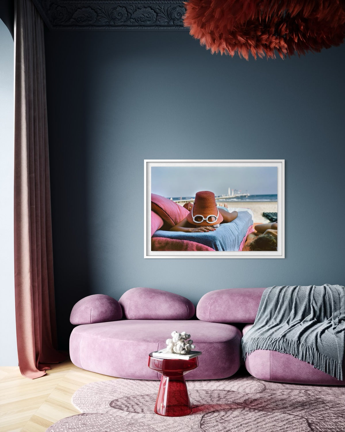 A framed art print depicting a cartoon character wearing a red bucket hat and sunglasses, lounging on a beach, is displayed on a dark slate blue wall above a modern purple sectional sofa in a stylishly decorated room.