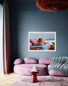 A framed art print depicting a cartoon character wearing a red bucket hat and sunglasses, lounging on a beach, is displayed on a dark slate blue wall above a modern purple sectional sofa in a stylishly decorated room.