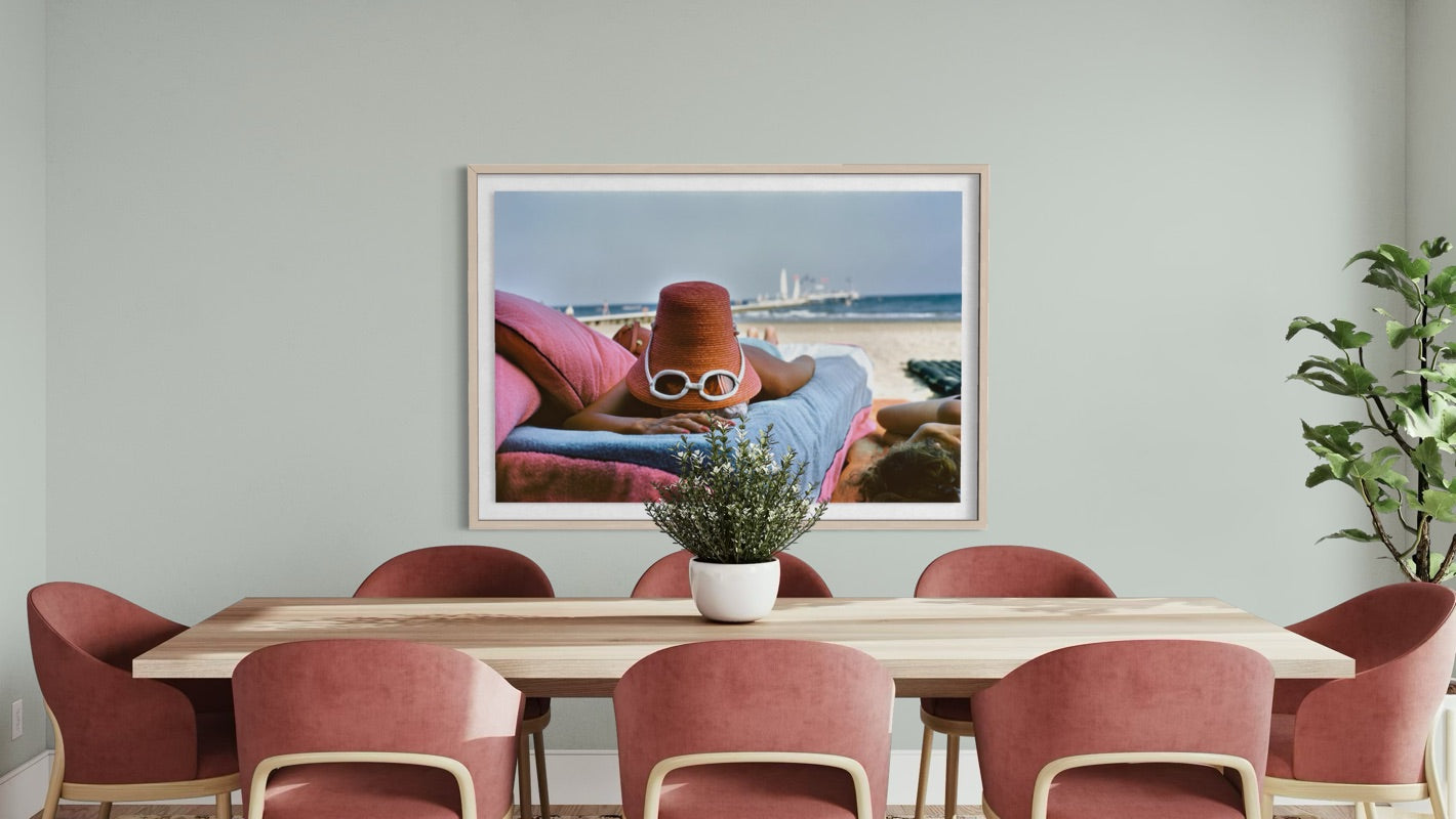 A framed art print depicting a woman lounging on a beach lounge chair by the ocean, hung on a light gray wall above a wooden dining table with pink chairs.