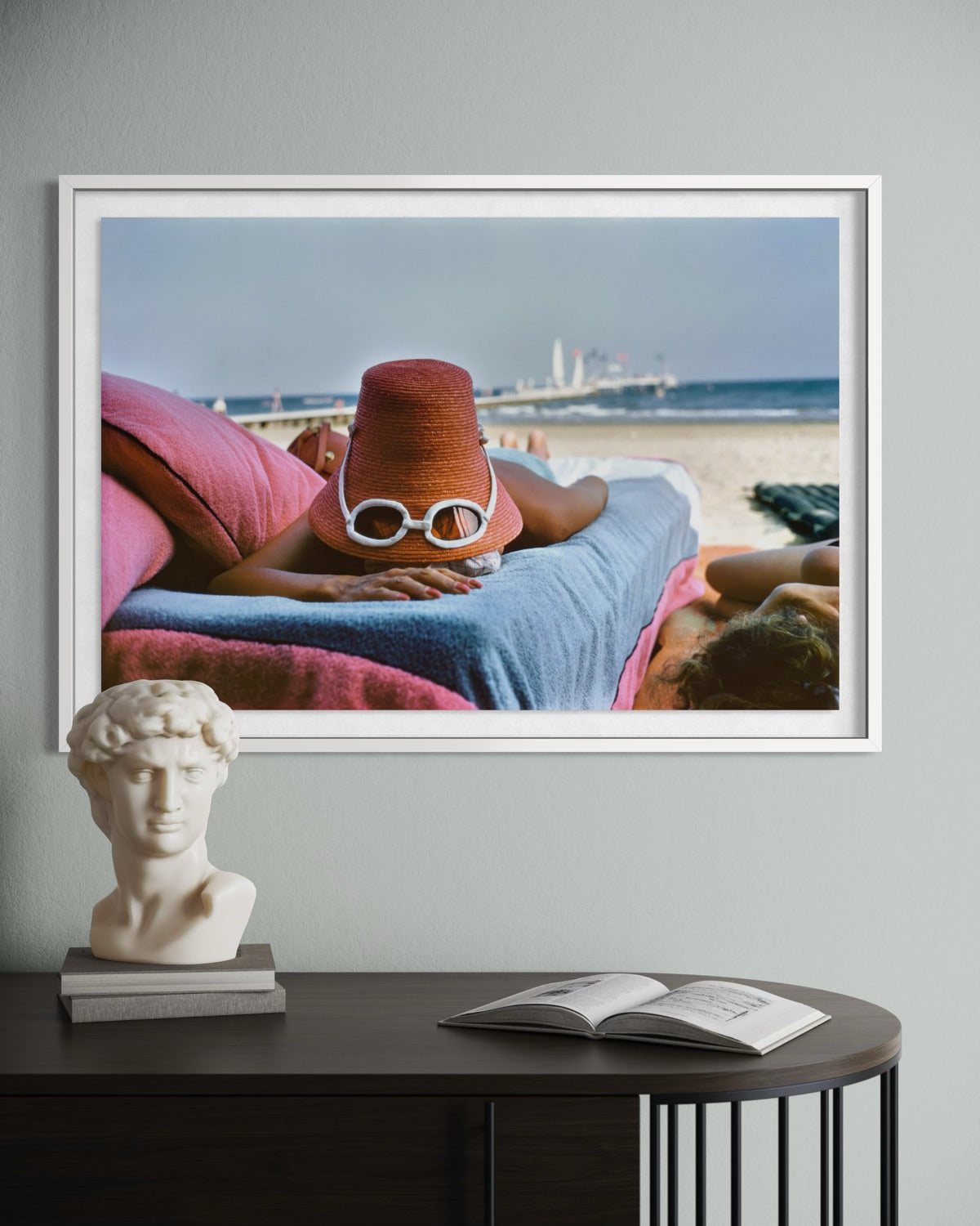 A framed art print depicting a woman relaxing on a beach lounge chair with a red bucket hat and white sunglasses, displayed on a light gray wall above a dark wooden console table with a white classical bust sculpture and an open book.