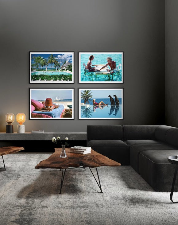 A modern living room setting featuring a four-panel framed wall art set depicting tropical beach and poolside scenes, mounted on a dark gray wall above a contemporary dark gray sectional sofa and a rustic wood coffee table.