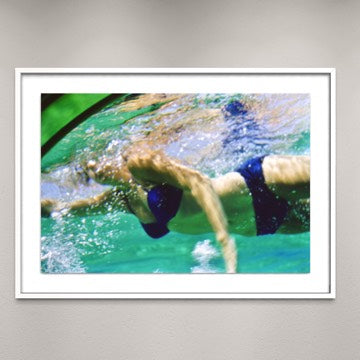 A framed photographic print of an underwater swimmer in a pool, shown hanging on a light gray wall. The print features a swimmer in dark blue swim trunks moving through turquoise water, enclosed in a white frame.