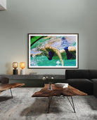 A framed art print depicting a swimmer in turquoise water, displayed on a gray wall in a modern living room setting above a wooden coffee table and dark sofa.