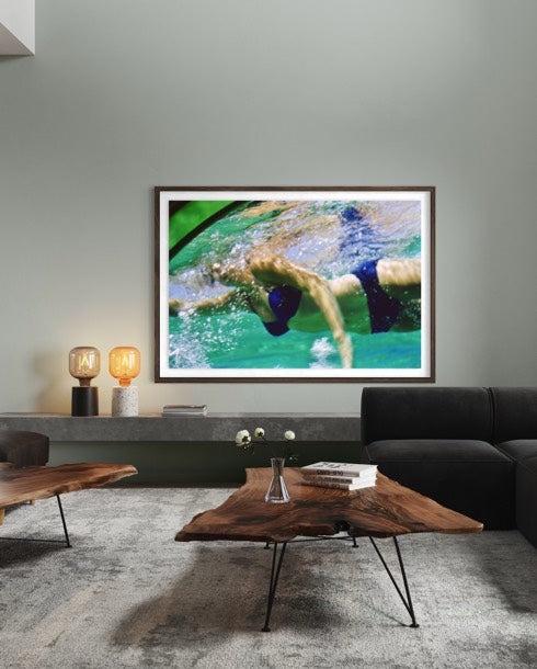 A framed art print depicting a swimmer in turquoise water, displayed on a gray wall in a modern living room setting above a wooden coffee table and dark sofa.