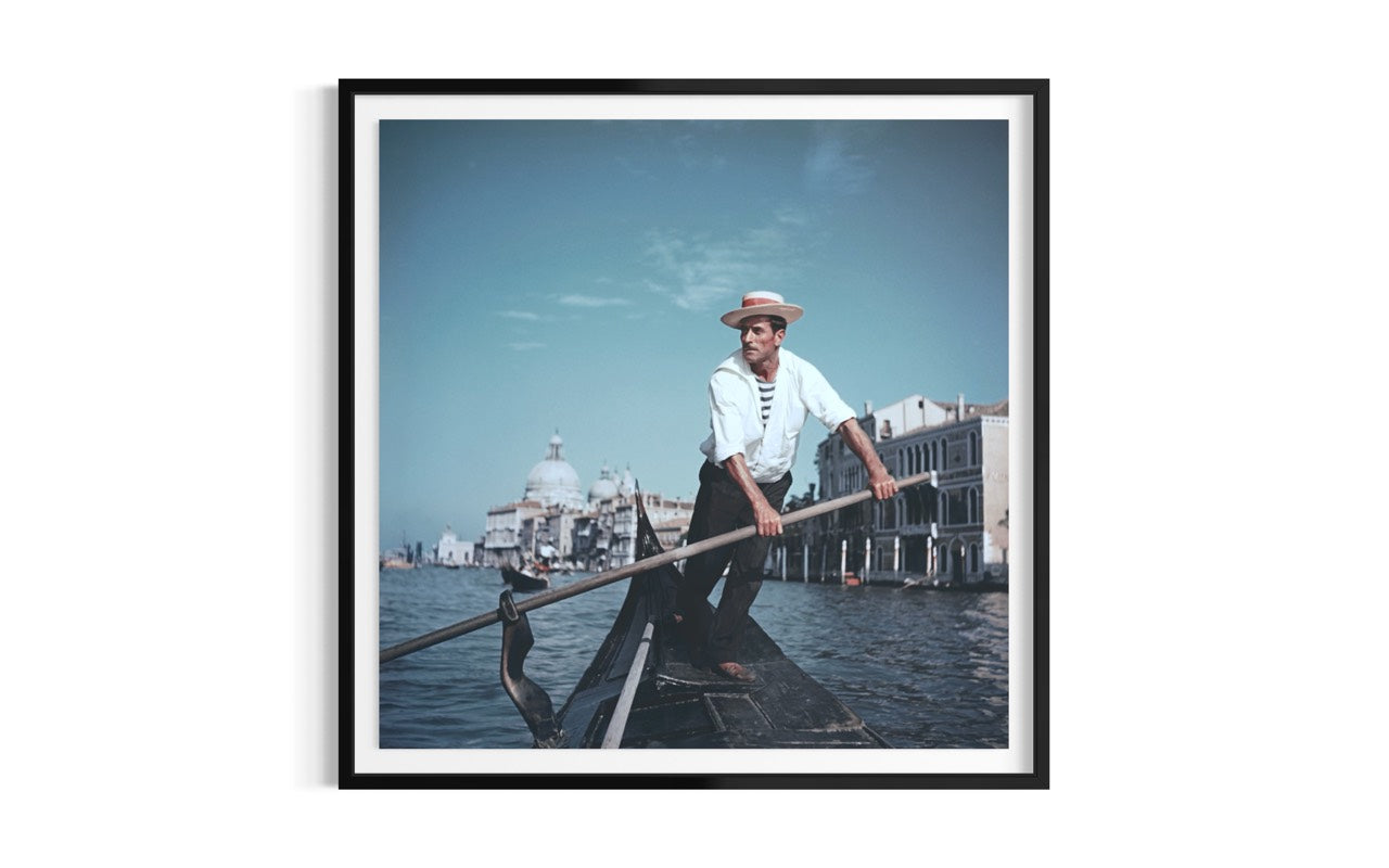 A framed art print featuring a vintage photograph of a gondolier in Venice, shown against a clean white background. The Picturalist Wall Art and Photography for Designed Interiors. Slim Aarons Collection, Venice. Getty Images Gallery. 