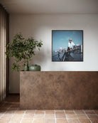 A framed art print depicting a gondolier in Venice on a canal, displayed on a neutral wall above a textured brown counter in a modern interior setting. The Picturalist Wall Art and Photography for Designed Interiors. Slim Aarons Collection, Venice. Getty Images Gallery. 