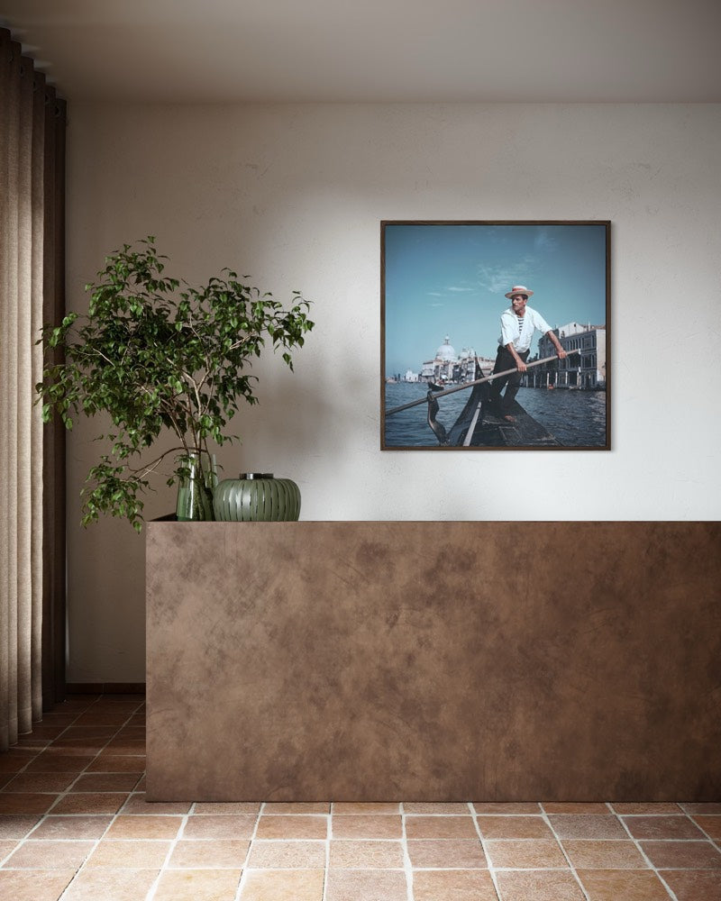 A framed art print depicting a gondolier in Venice on a canal, displayed on a neutral wall above a textured brown counter in a modern interior setting. The Picturalist Wall Art and Photography for Designed Interiors. Slim Aarons Collection, Venice. Getty Images Gallery. 