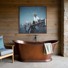 A framed art print depicting a gondolier in a gondola in Venice, hung on a wooden plank wall above a freestanding copper bathtub. The Picturalist Wall Art and Photography for Designed Interiors. Slim Aarons Collection, Venice. Getty Images Gallery. 