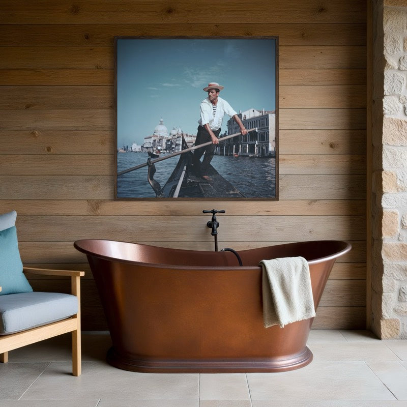 A framed art print depicting a gondolier in a gondola in Venice, hung on a wooden plank wall above a freestanding copper bathtub. The Picturalist Wall Art and Photography for Designed Interiors. Slim Aarons Collection, Venice. Getty Images Gallery. 