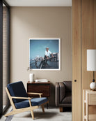 A framed art print depicting a man rowing a boat on a river is displayed on a beige wall in a modern, stylishly decorated room with a blue armchair, wooden furniture, and a lamp. The Picturalist Wall Art and Photography for Designed Interiors. Slim Aarons Collection, Venice. Travel. Getty Images Gallery. 