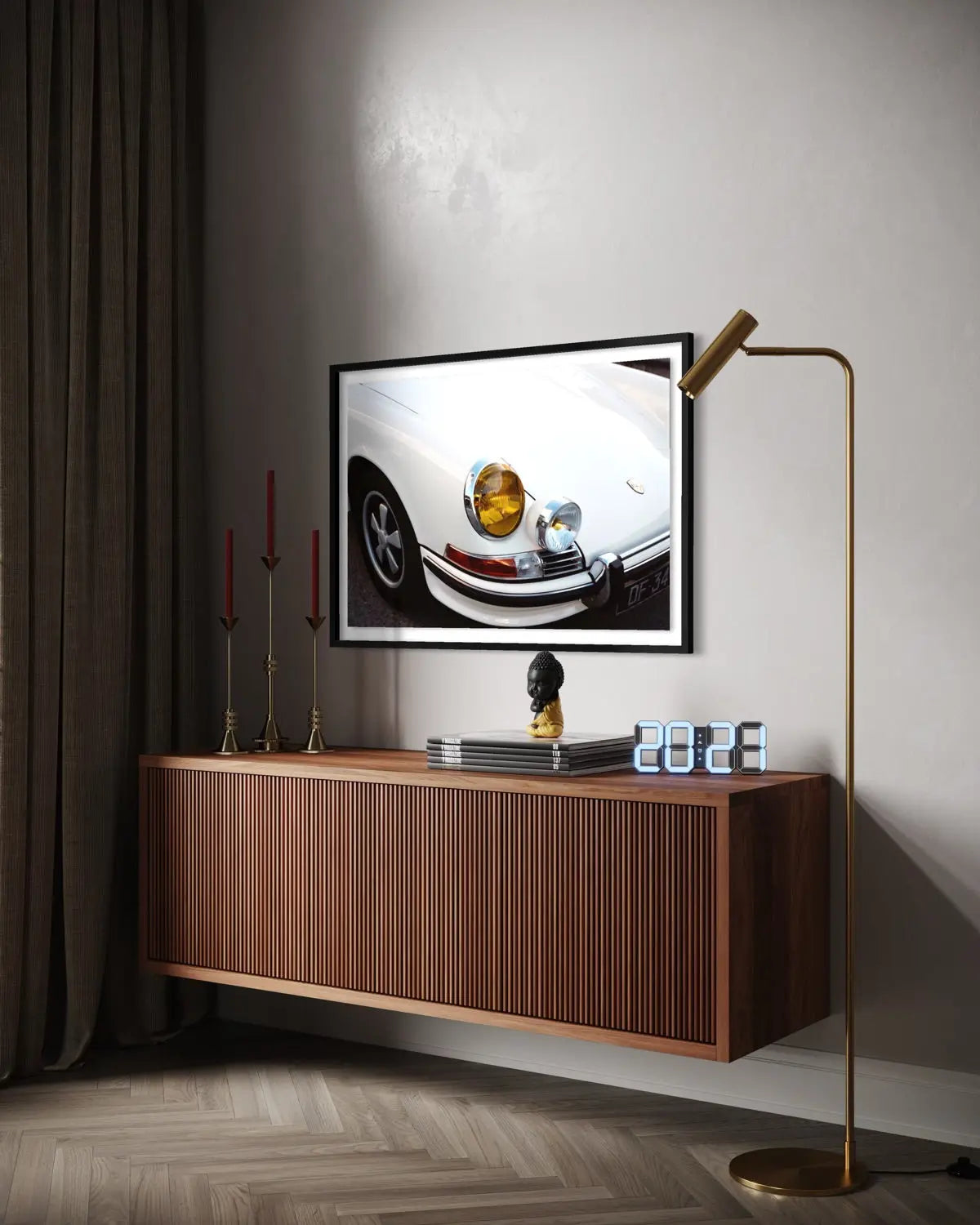 A framed wall art print depicting a close-up of a white car's front end with a gold headlight, displayed above a floating wooden console in a modern room setting with a gold floor lamp and dark curtains.