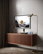 A framed wall art print depicting a close-up of a white car's front end with a gold headlight, displayed above a floating wooden console in a modern room setting with a gold floor lamp and dark curtains.