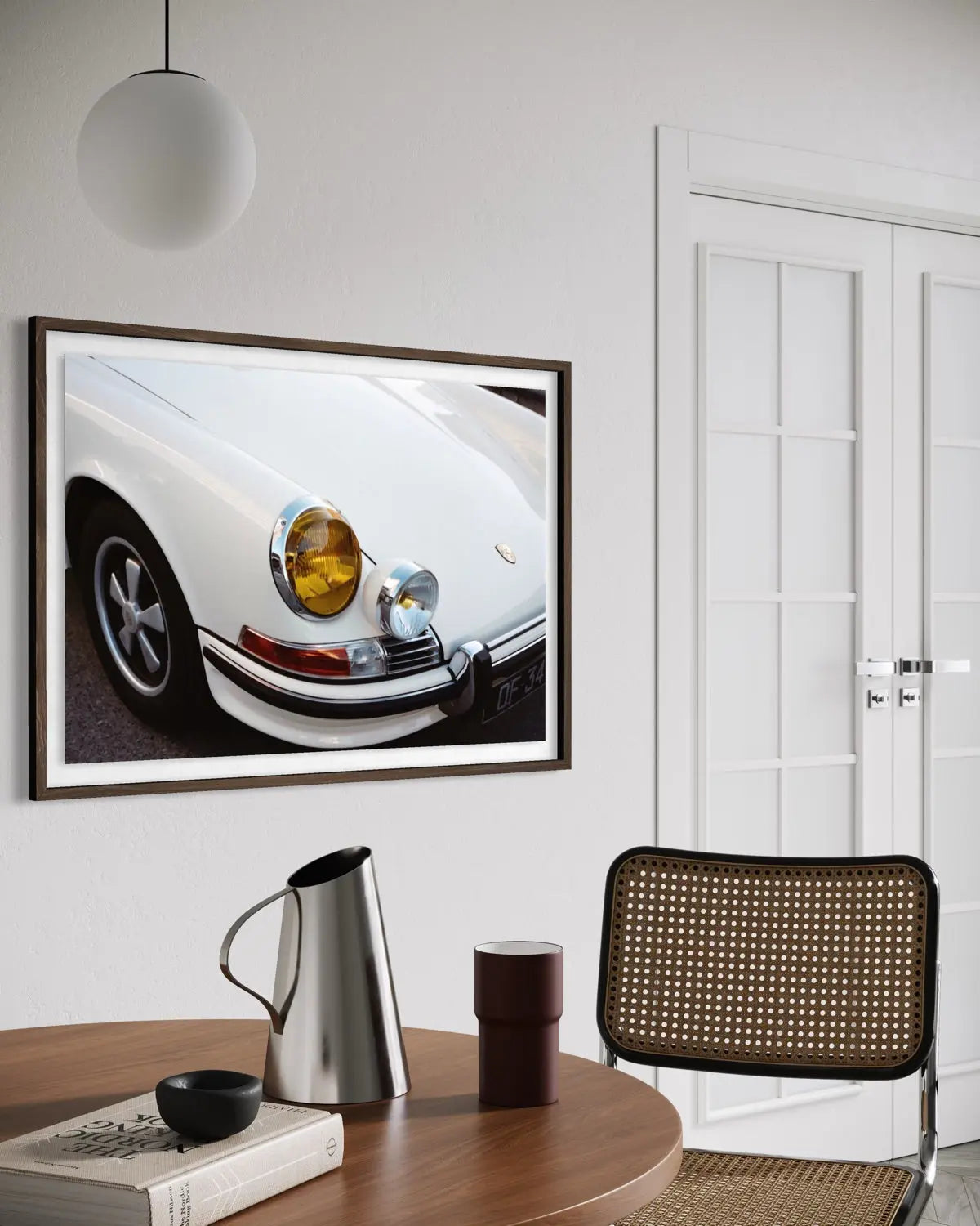 A framed art print of a white classic Porsche car hanging on a white wall in a modern dining room setting with a wooden table, chair, and coffee accessories.