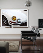 A framed wall art print showing a close-up photograph of a white classic car, displayed in a modern living room setting above a white console table with two black chairs and a speaker.